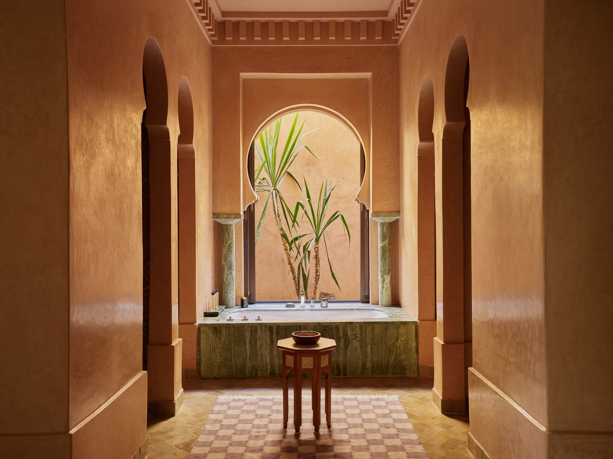 Amanjena, Africa — Ochre-walled hammam with arched doorway, sunlit bath basin, green-glazed tile work, potted palms, and carved wooden columns.