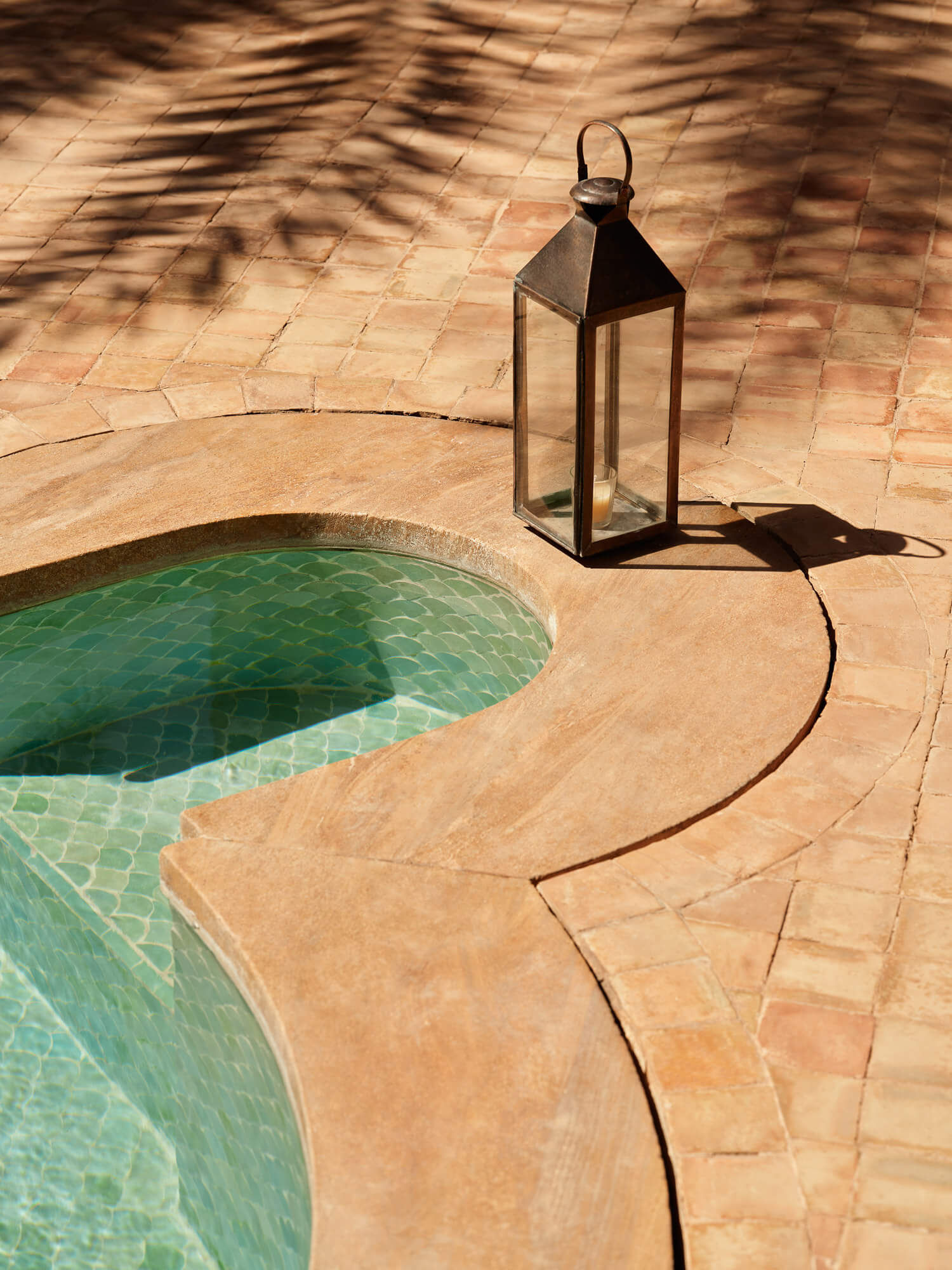 Amanjena, Africa — Curved stone terrace with turquoise pool edge, metal lantern casting shadows on warm tile paving, minimalist luxury courtyard design.