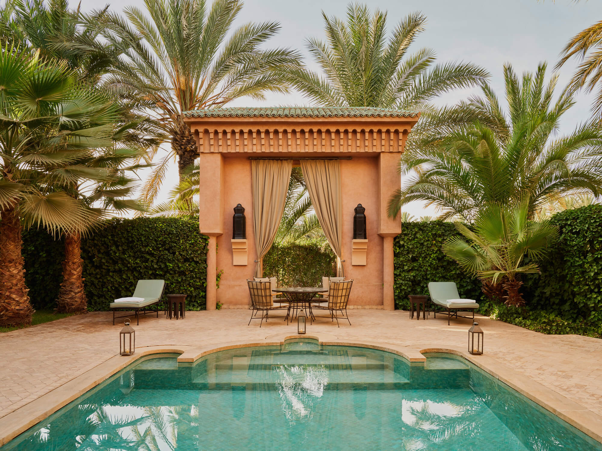 Amanjena, Morocco — Terracotta pavilion with draped curtains overlooking turquoise pool, flanked by palm trees and loungers on ochre stone terrace.