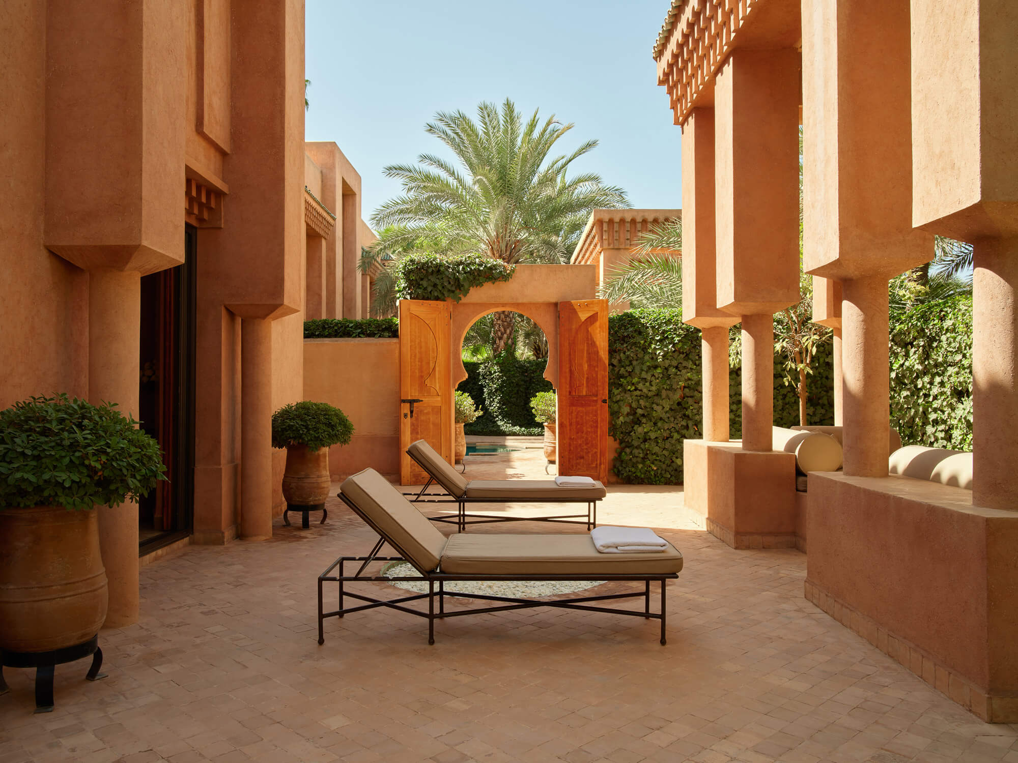 Amanjena, Africa — Terracotta courtyard with loungers, arched doorway, palm trees, and ochre-rendered walls under clear sky.