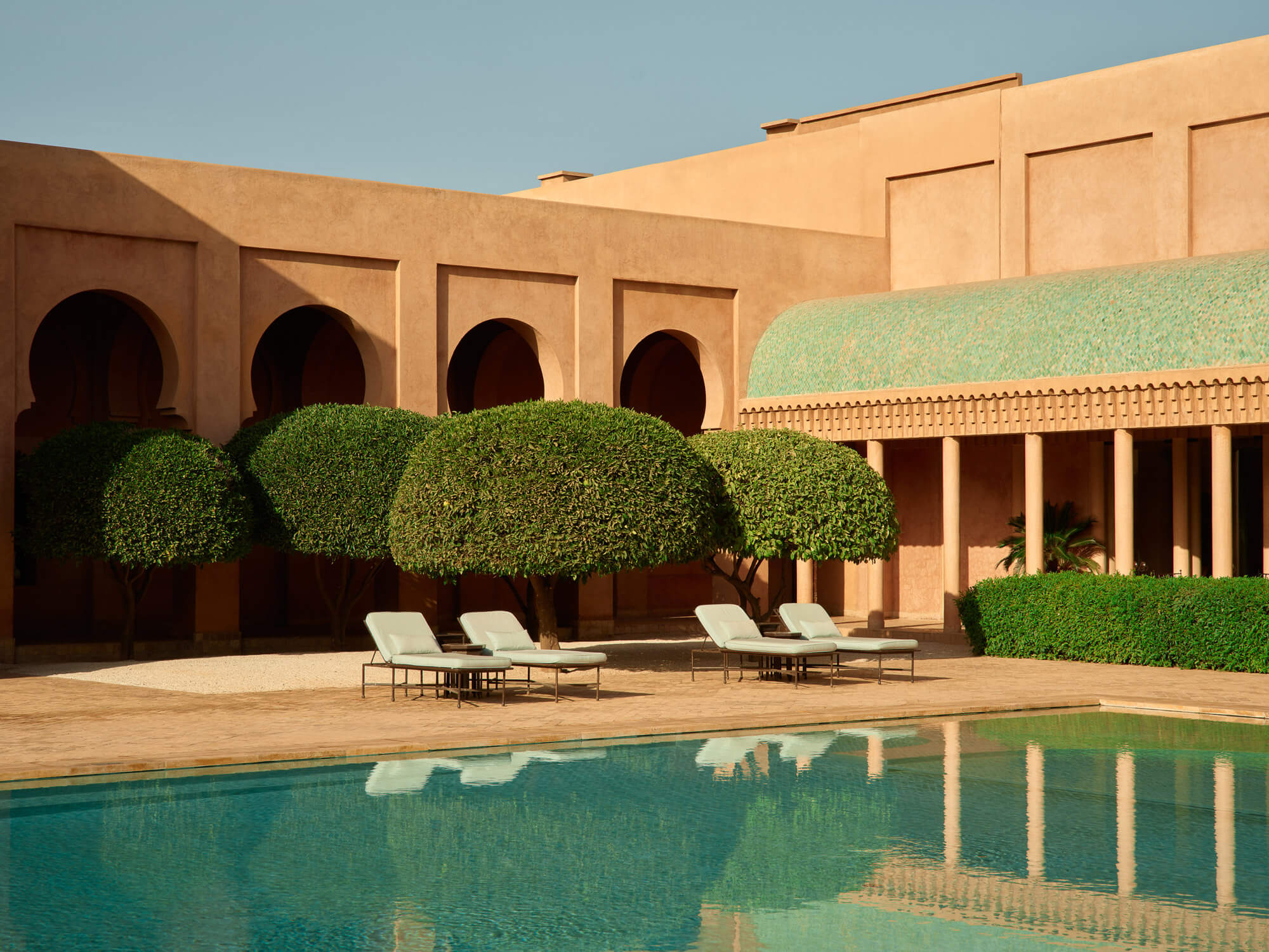 Amanjena, Africa — Ochre-walled courtyard with arched alcoves, manicured topiaries, stone loungers, and turquoise pool reflecting modernist minimalist architecture under clear sky.