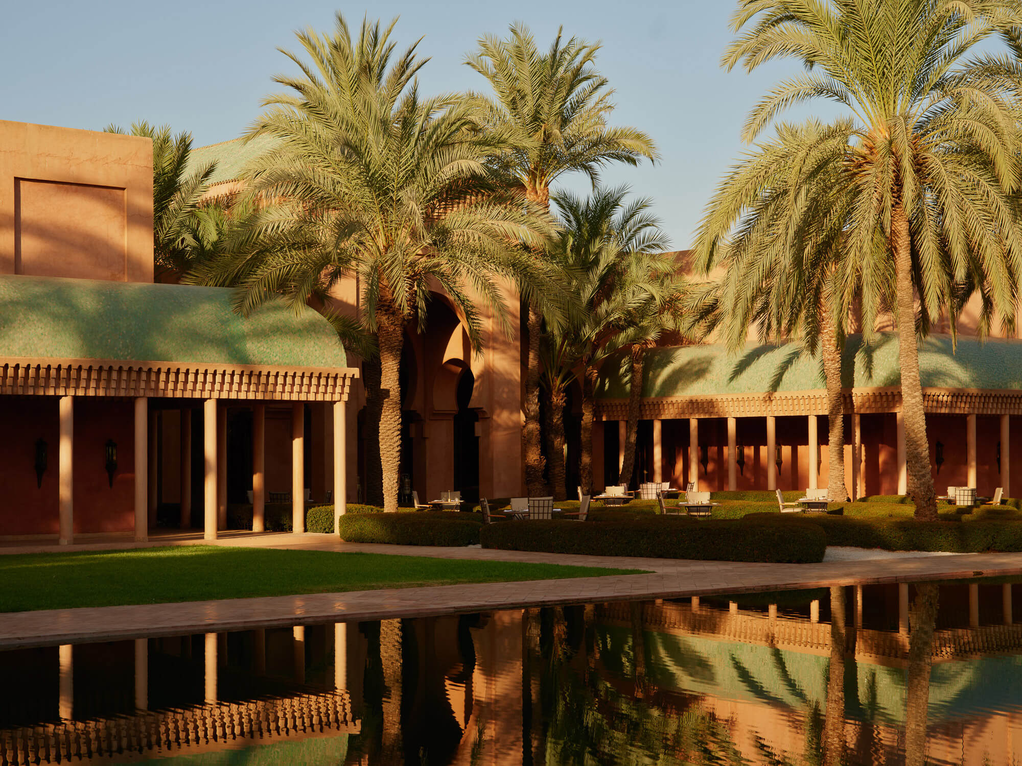 Amanjena, Marrakech, Morocco — Terracotta courtyard with colonnade, palm trees, reflecting pool, and desert modernist architecture in golden light.