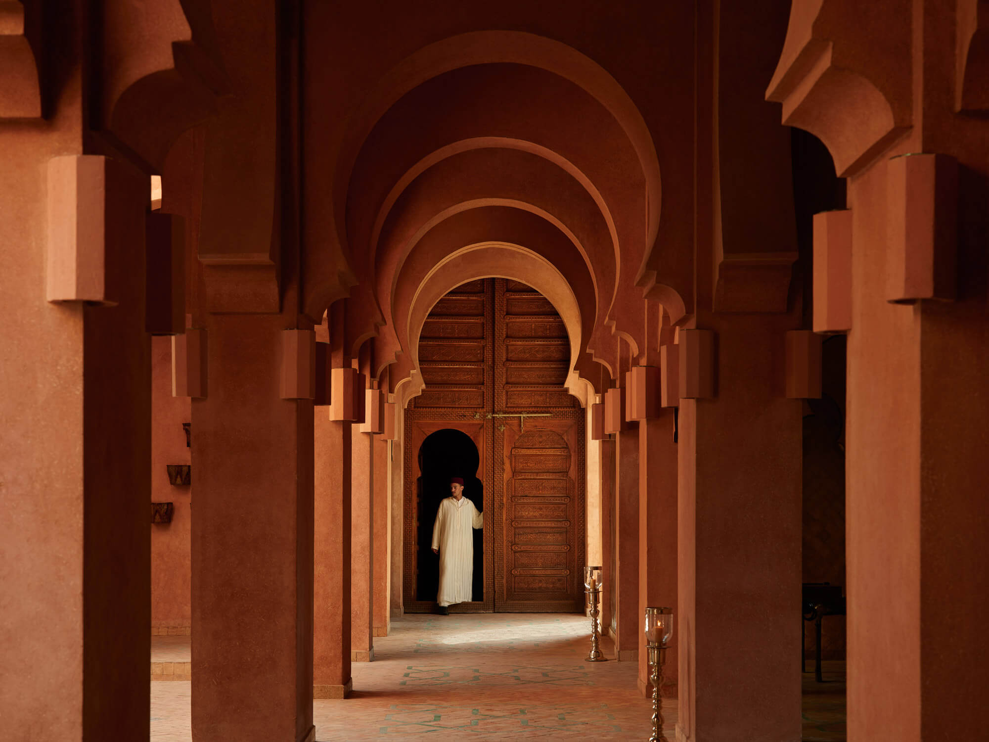 Amanjena, Marrakech, Morocco — Terracotta arches and columns frame wooden door in candlelit Islamic corridor, minimalist luxury architecture.