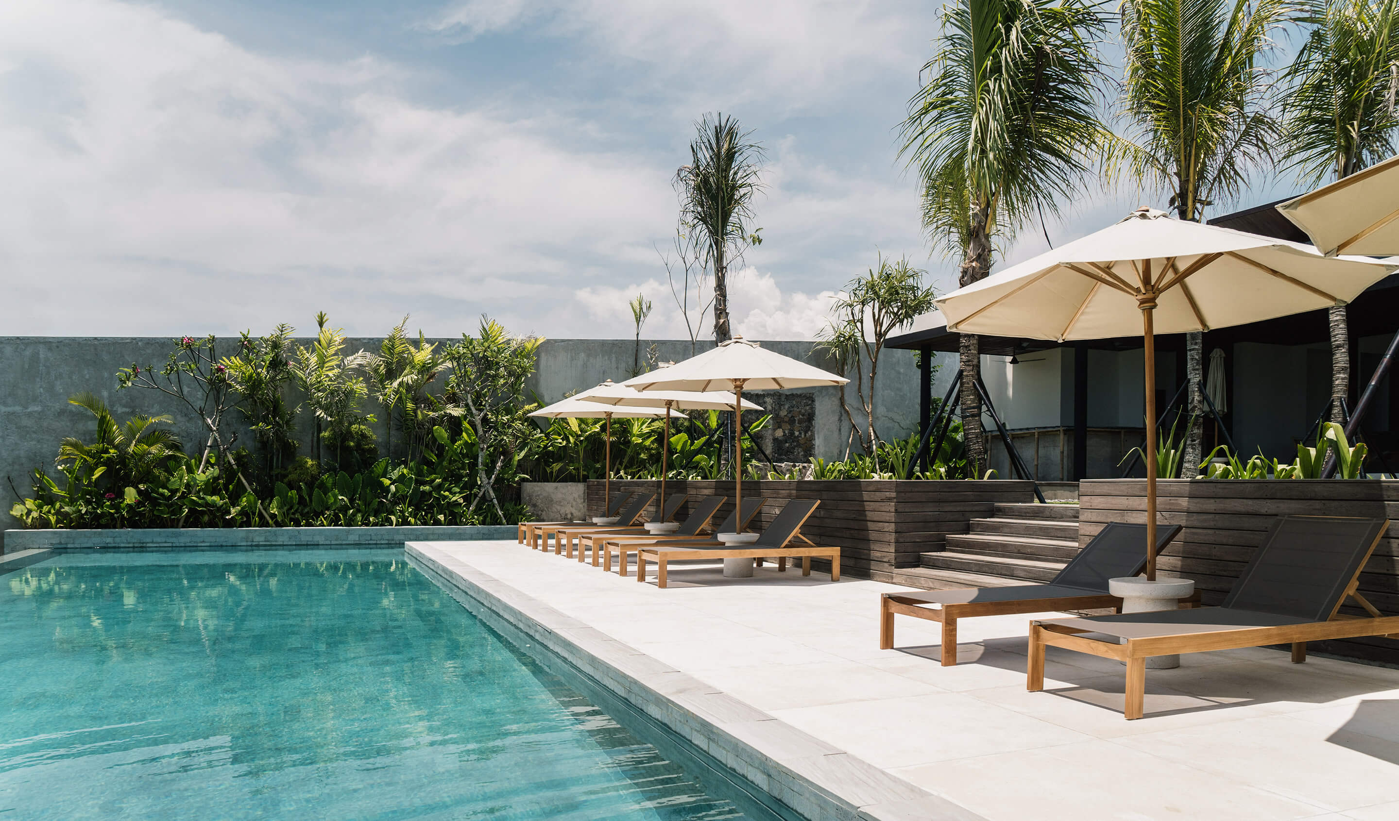 Somewhere Lombok, Indonesia — Poolside lounge with teak sunbeds, canvas umbrellas, tropical palms, and modern concrete architecture under bright daylight.