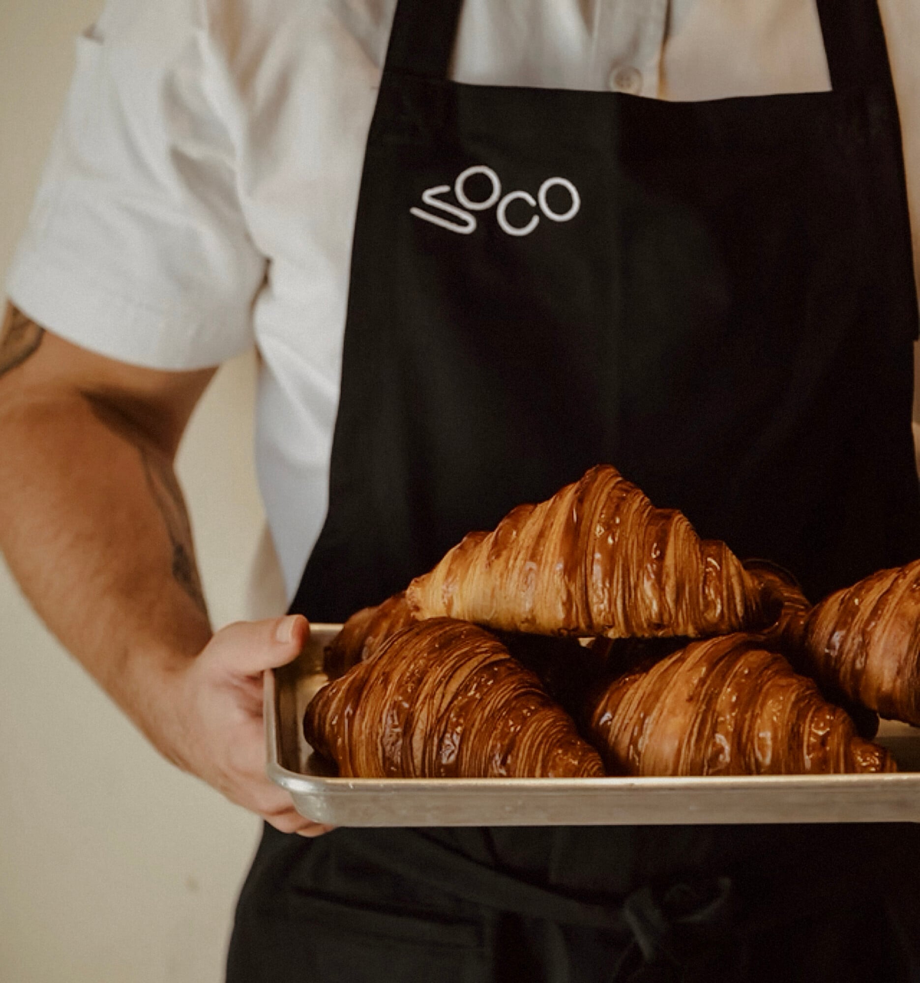 Soco, Mexico — Baker holding tray of chocolate croissants in black apron, artisanal pastries with glossy laminated layers in natural light.