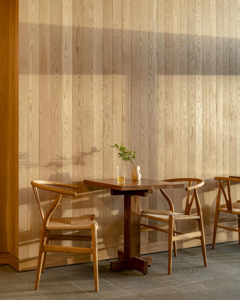 Piaule, North America — Mid-century wooden chairs and round pedestal table bathed in afternoon sunlight against vertical wood-paneled wall, minimalist hospitality interior.