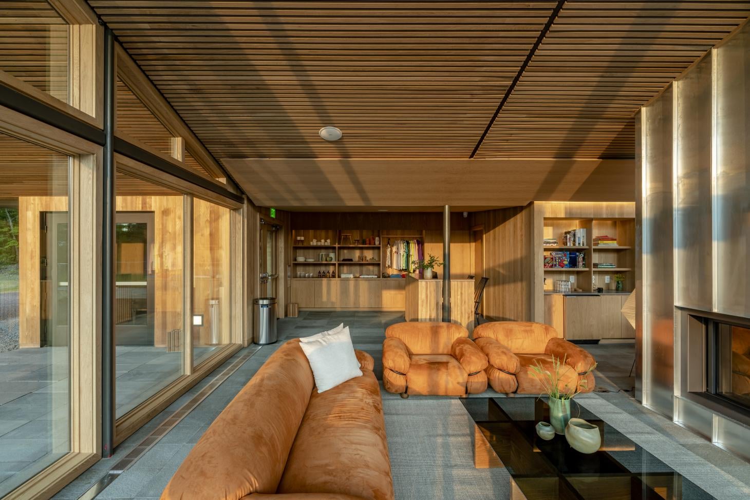 Piaule, North America — Modern wooden interior with cognac leather seating, linear ceiling details, floor-to-ceiling glazing, and built-in shelving in warm natural light.