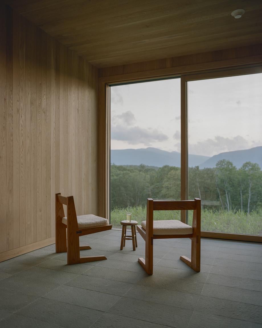 Piaule, North America — Minimalist wooden villa interior with mid-century chairs facing floor-to-ceiling windows overlooking forested mountains and valley landscape.
