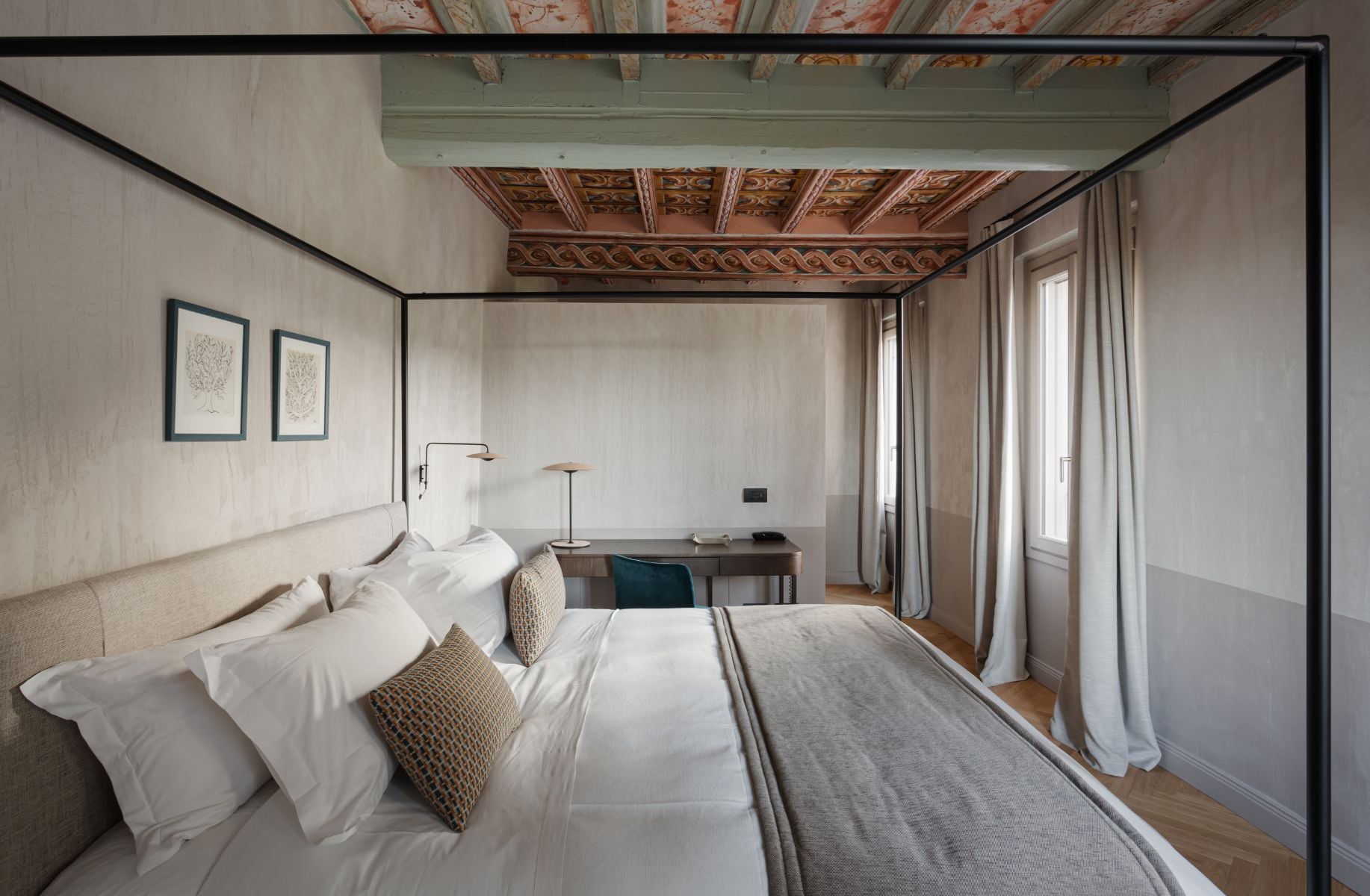 Roncolo 1888, Europe — Canopy bed bedroom with ornate wooden ceiling beams, minimalist furnishings, natural light from French doors, and contemporary desk workspace.