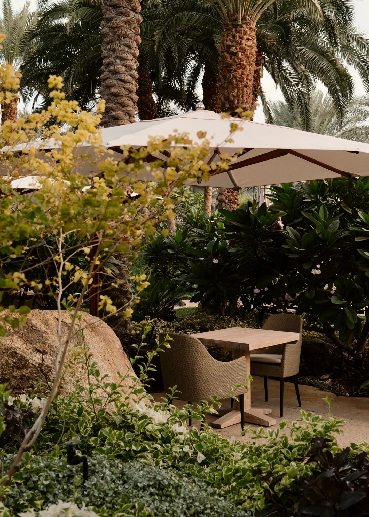 Sensei Porcupine Creek, North America — Shaded outdoor seating beneath cantilever umbrella amid palm trees, flowering shrubs, and natural stone in minimalist luxury garden design.