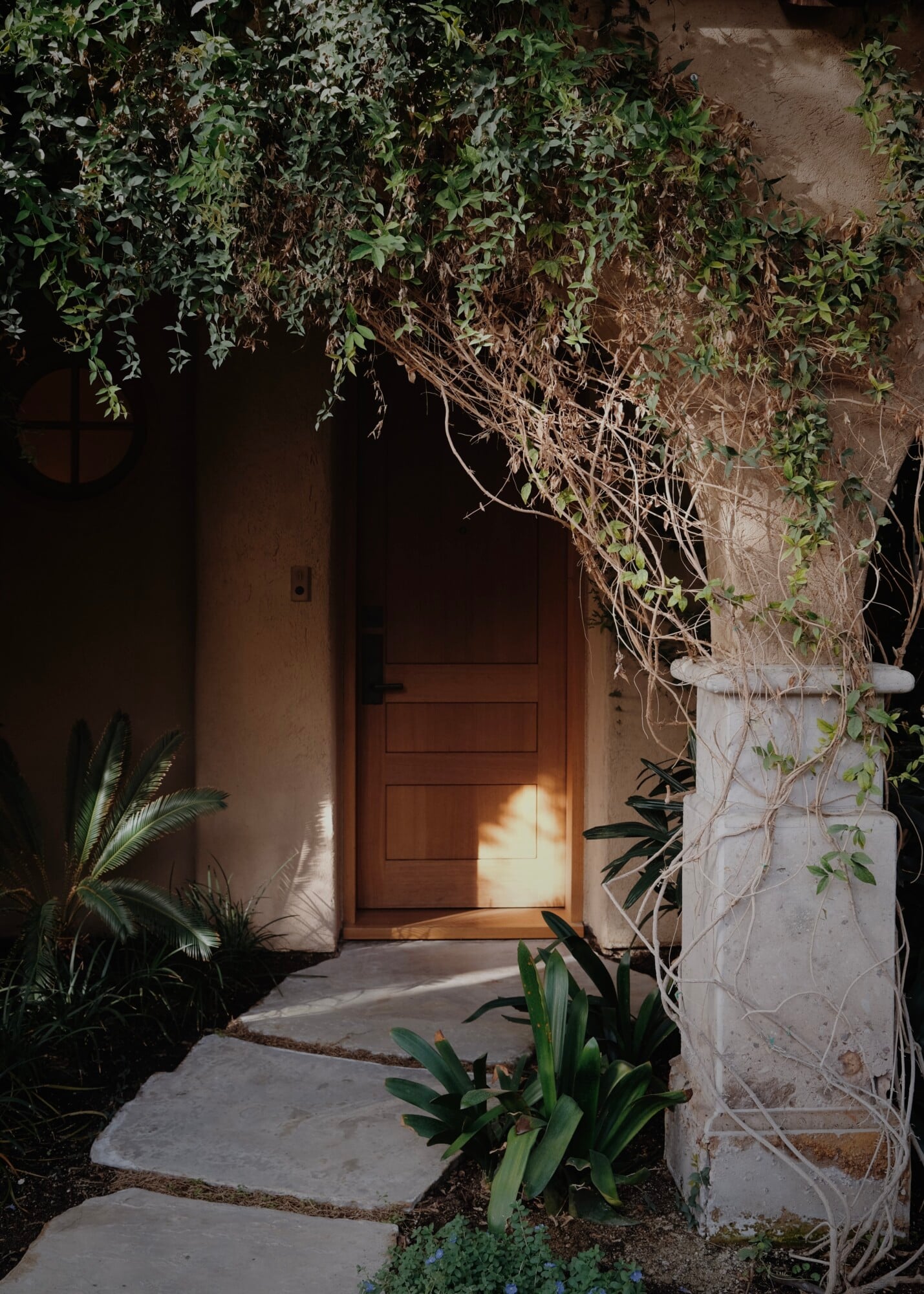 Sensei Porcupine Creek, North America — Illuminated wooden entry door framed by trailing vines, stone column, and lush desert plantings along stepped stone pathway at dusk.