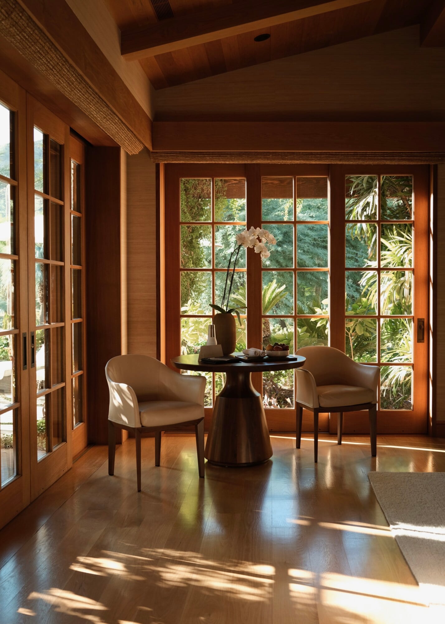 Sensei Porcupine Creek, North America — Wooden sunroom with grid-pane doors, paired leather chairs, pedestal table, orchid arrangement, forest views, warm natural light.