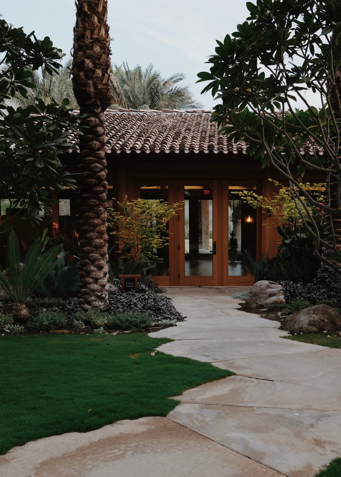 Sensei Porcupine Creek, North America — Terracotta-roofed pavilion with wood doors framed by palm trees and desert landscaping along stone pathway.