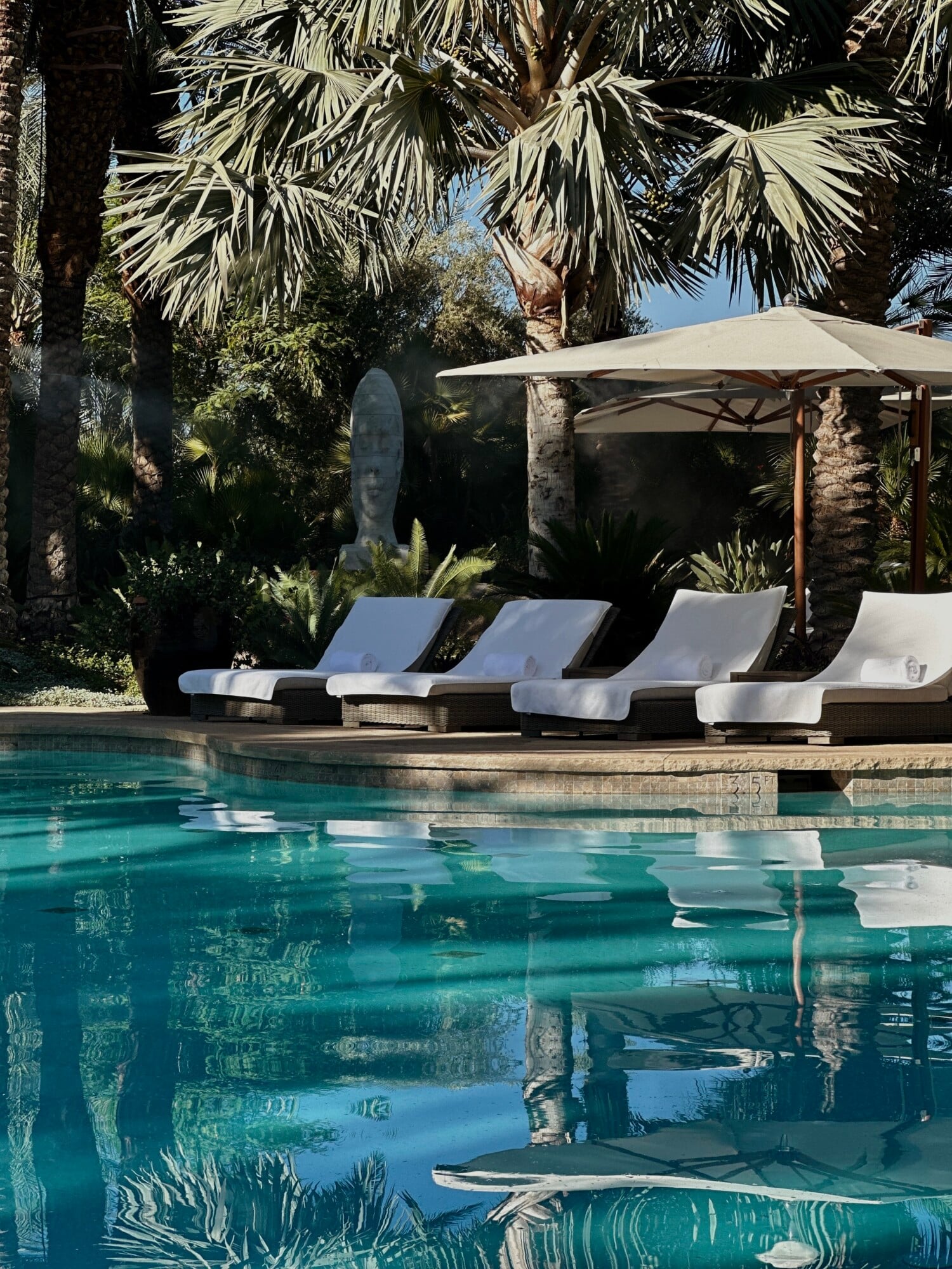 Sensei Porcupine Creek, North America — Poolside lounge chairs with white cushions beneath a cantilever umbrella, surrounded by palm trees and desert landscaping with mountain views.
