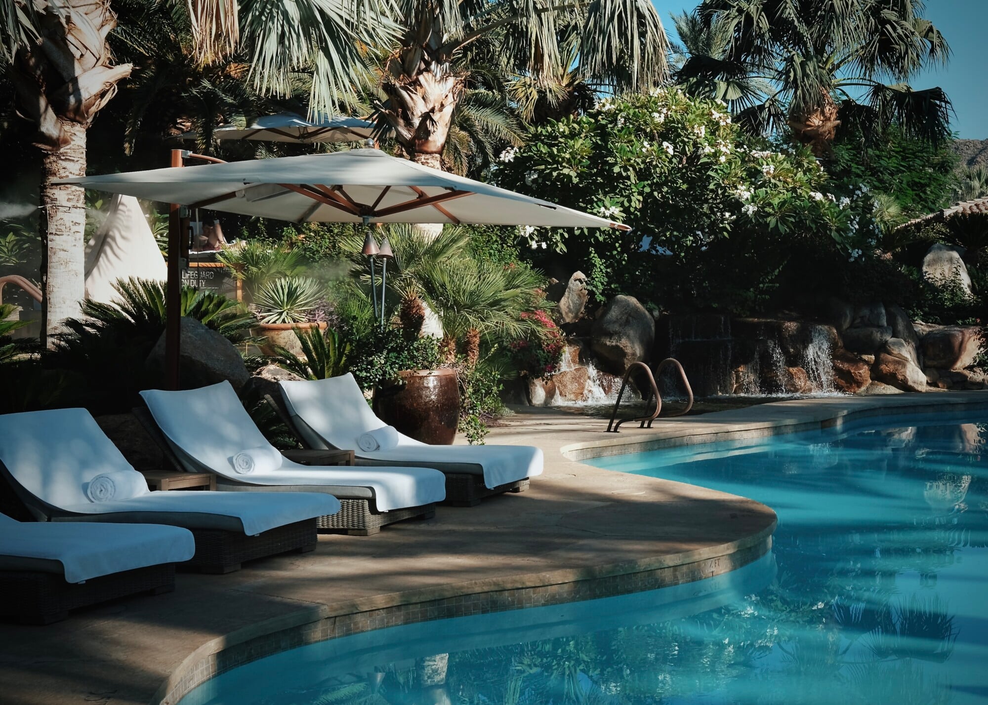 Sensei Porcupine Creek, North America — Poolside lounge chairs with white cushions beneath cantilever umbrella, desert landscaping with palms and succulents, architectural water feature.