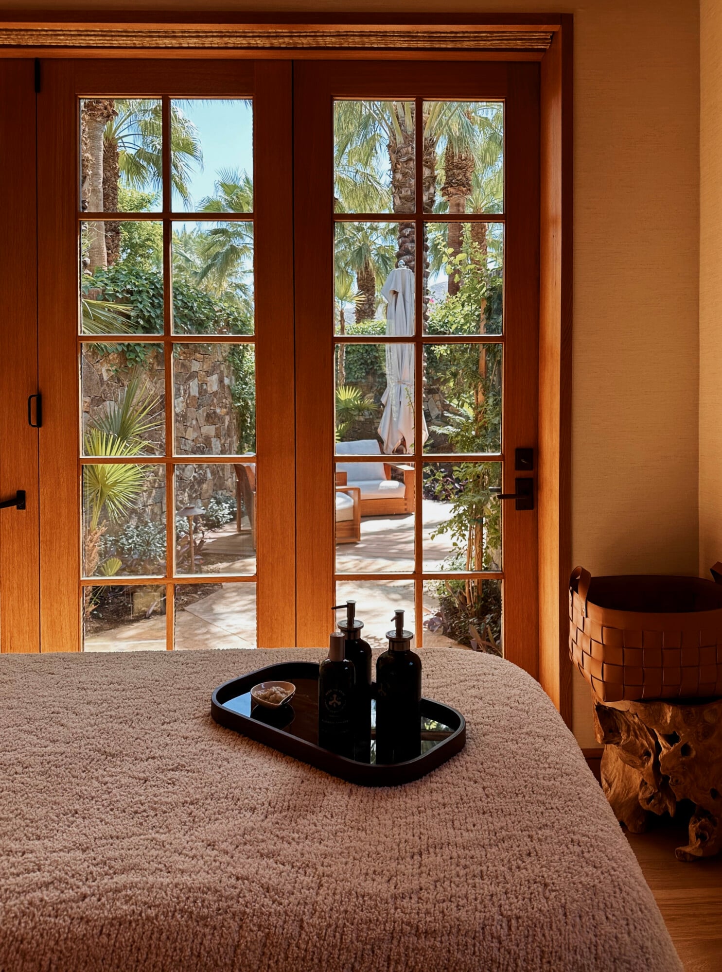 Sensei Spa, North America — Wooden French doors with grid panes opening onto tropical garden with palm trees, natural light flooding minimal interior with linen-draped table and black accessories.