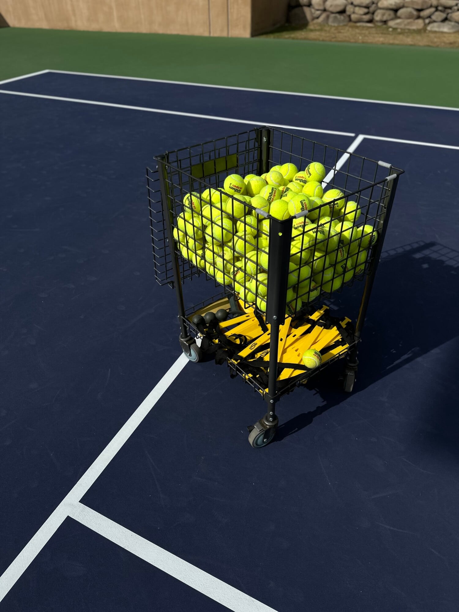 Sensei Porcupine Creek, North America — Tennis ball hopper on hard court with white boundary lines, morning light casting shadows across playing surface.