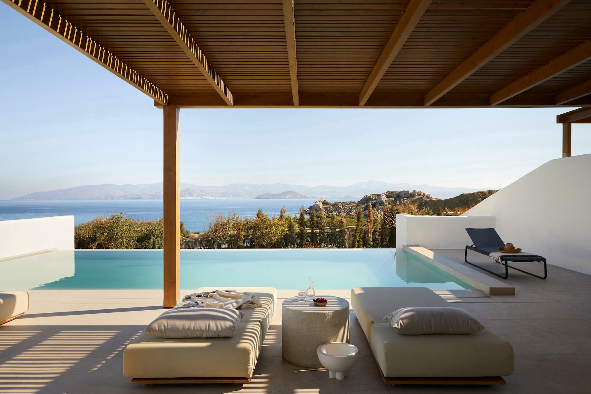 Pārocks, Europe — Modern waterfront terrace with infinity pool, wooden pergola, minimal furnishings, and expansive sea view across forested coastline at golden hour.
