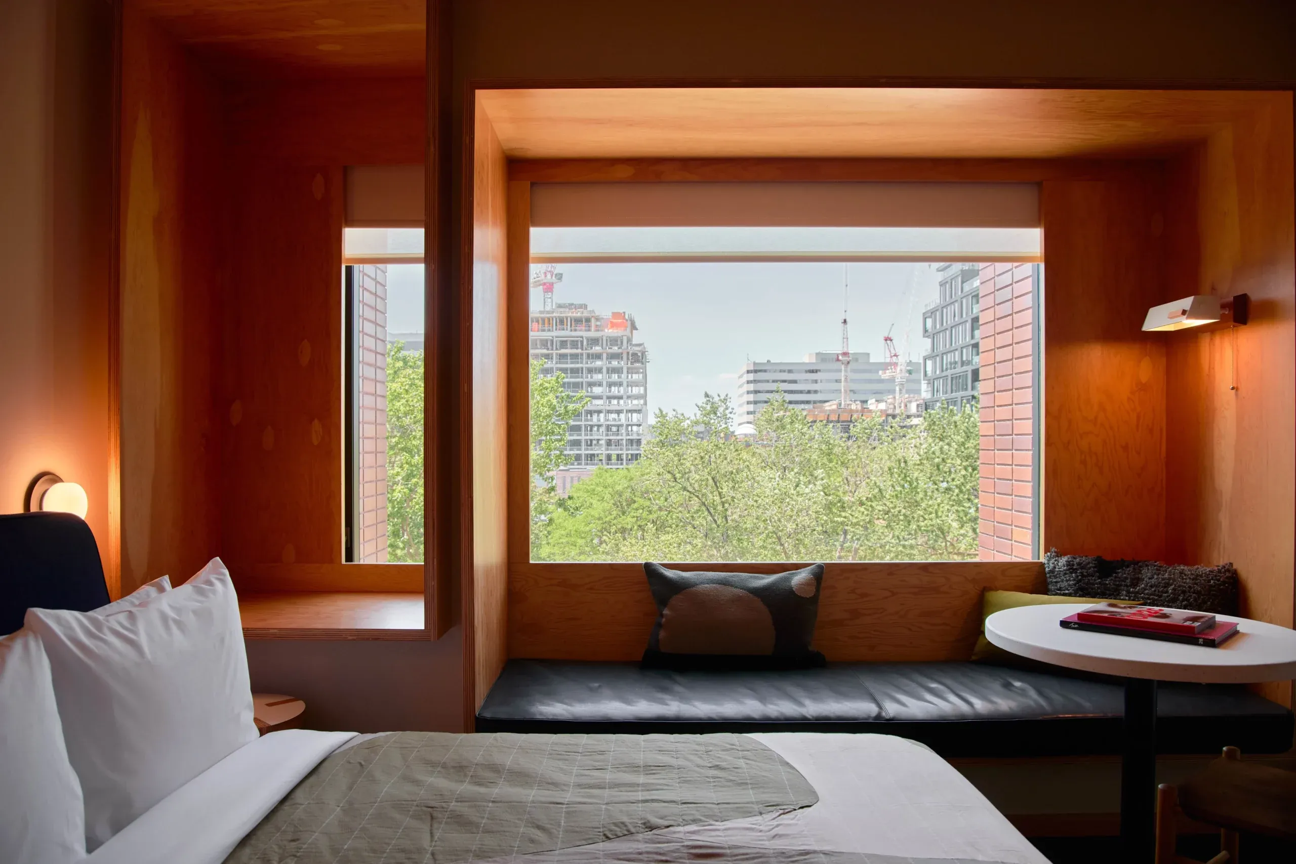 Ace Hotel Toronto, Canada — Warm wood-framed window seat overlooking urban skyline, minimal bed with white linens, black leather bench, natural light, modernist hospitality design.