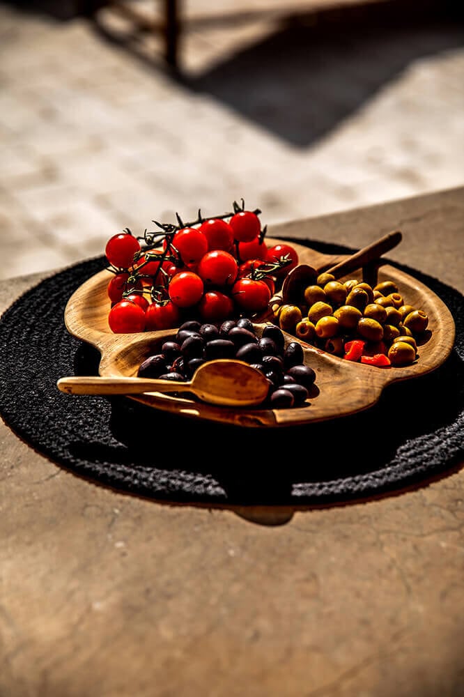 Es Bec d’Aguila — Es Bec d'Aguila, Balearic Islands, Spain — Wooden palette dish with cherry tomatoes, olives, and capers on black linen, sunlit Mediterranean styling.