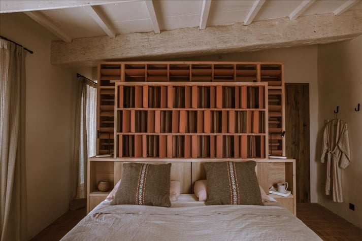 Son Blanc Farmhouse, Balearic Islands, Spain — Minimalist bedroom with custom wooden headboard bookshelf, natural linen bedding, exposed stone ceiling beams, warm neutral tones.