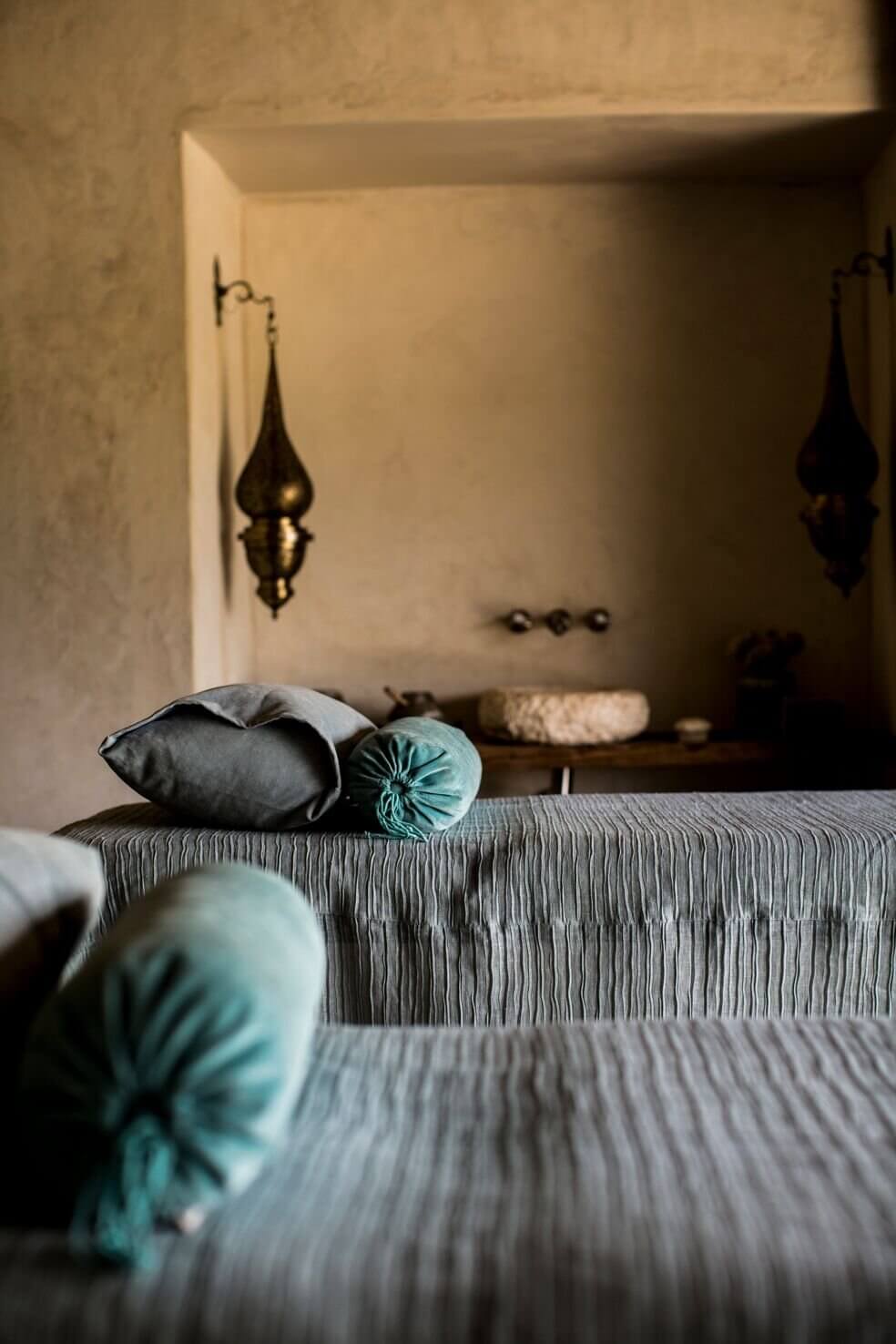 Arijiju Retreat, Africa — Minimalist bedroom with striped linen bedding, turquoise accent pillows, recessed alcove with brass lanterns, earthy neutral palette, warm natural light.
