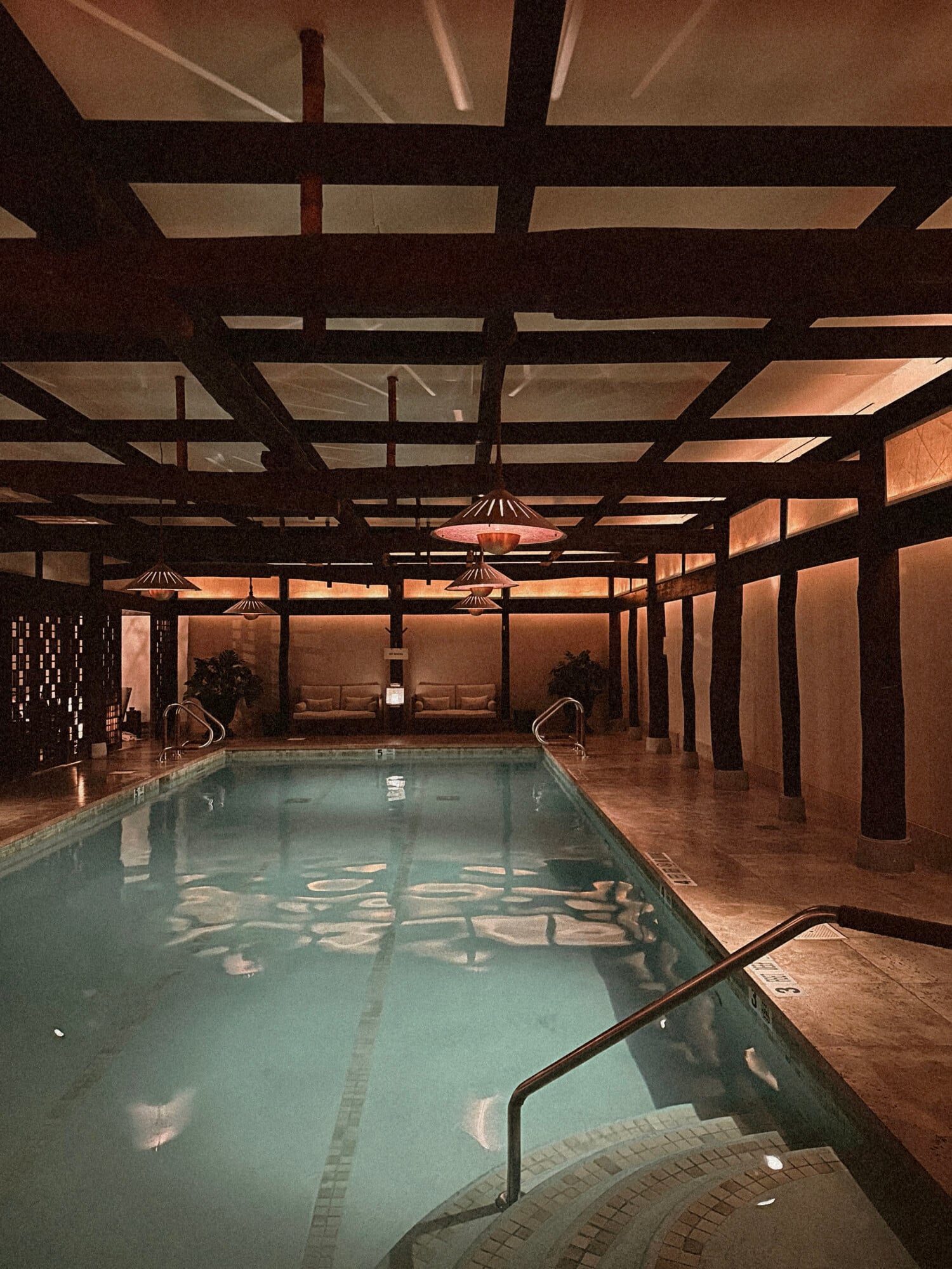 Shibui Spa, North America — Indoor lap pool with wooden beam ceiling, pendant lamps, and warm ambient lighting reflecting on calm water.