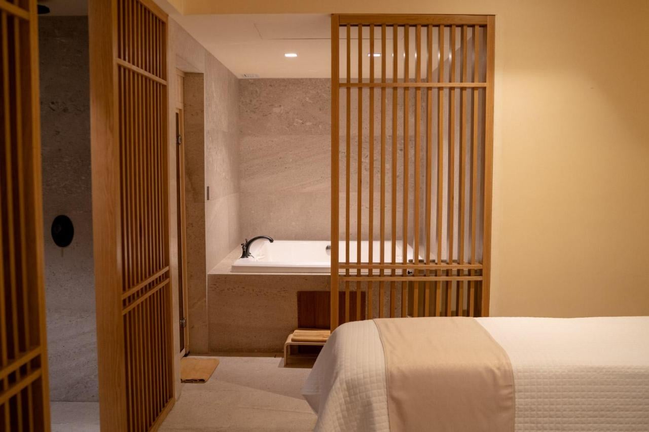 Esencia Spa, Mexico — Minimalist spa bedroom with wooden slat room divider, sunken soaking tub, natural stone walls, and warm ambient lighting.
