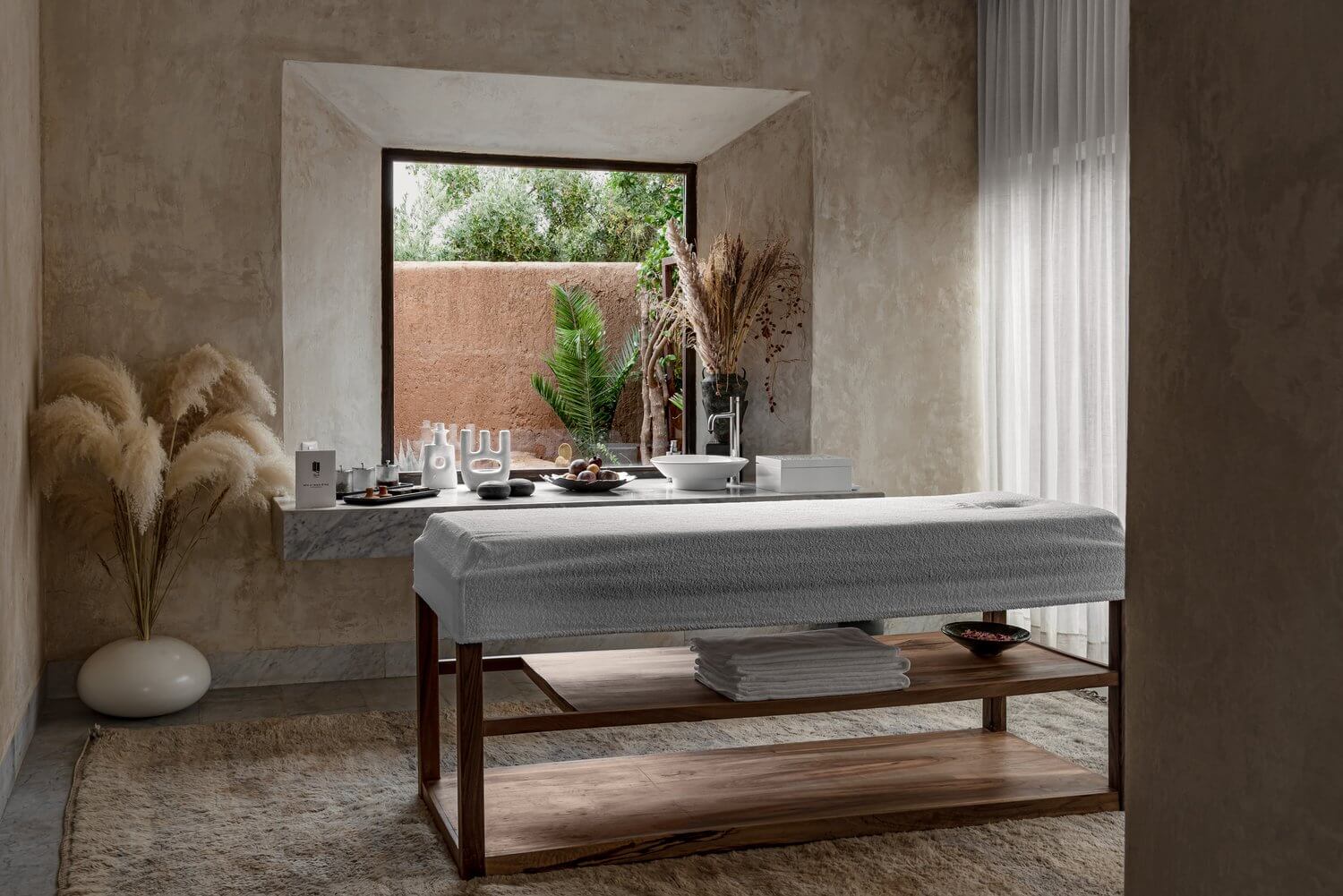 Spa & Yoga Pavillon, Africa — Minimalist spa bathroom with concrete walls, marble vanity, wooden massage table, pampas grass, and large window overlooking courtyard garden.
