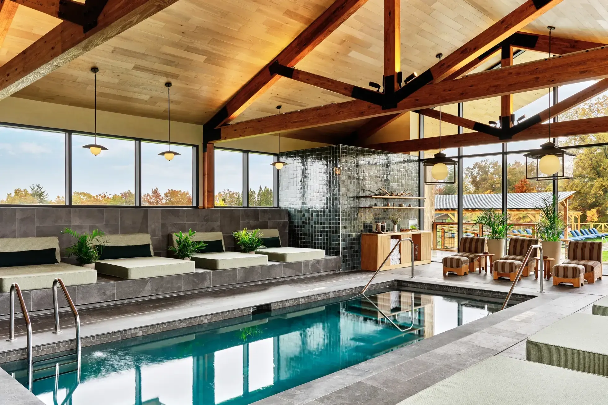 Wildflower Farms, North America — Indoor lap pool with turquoise water, exposed wood beam ceiling, floor-to-ceiling windows, gray tile surround, lounge seating with striped cushions, pendant lighting.