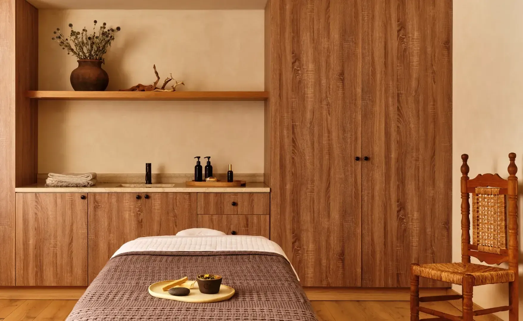 Thistle Spa, North America — Minimalist spa treatment room with warm wood cabinetry, floating shelf, cream walls, massage bed with grey linens, and natural light.