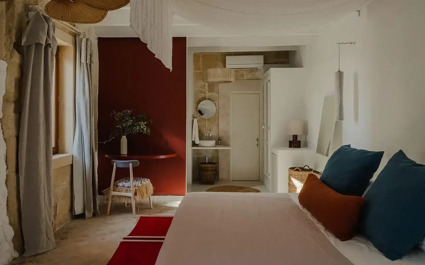 Amagatay, Balearic Islands, Spain — Minimalist hotel bedroom with deep red accent wall, terracotta bed frame, teal pillows, ensuite bathroom, natural light, Mediterranean design aesthetic.