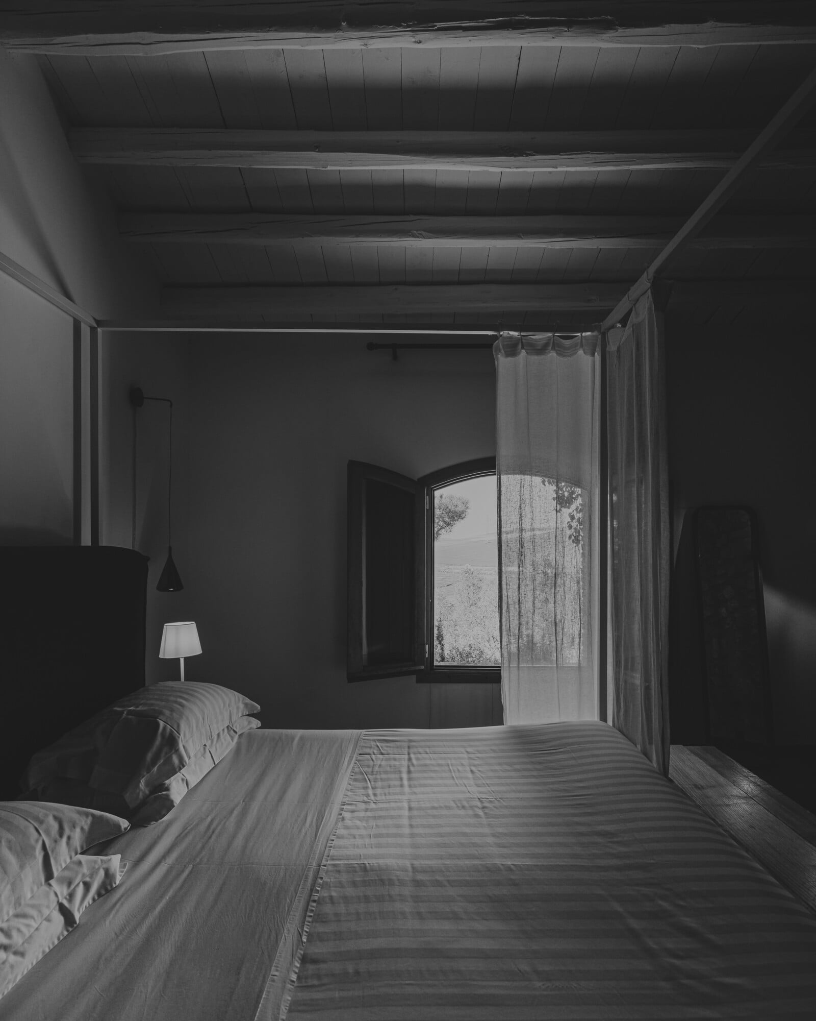 Susafa, Italy — Minimalist bedroom with canopy bed, sheer curtains, wooden ceiling beams, and open doors overlooking landscape in soft natural light.
