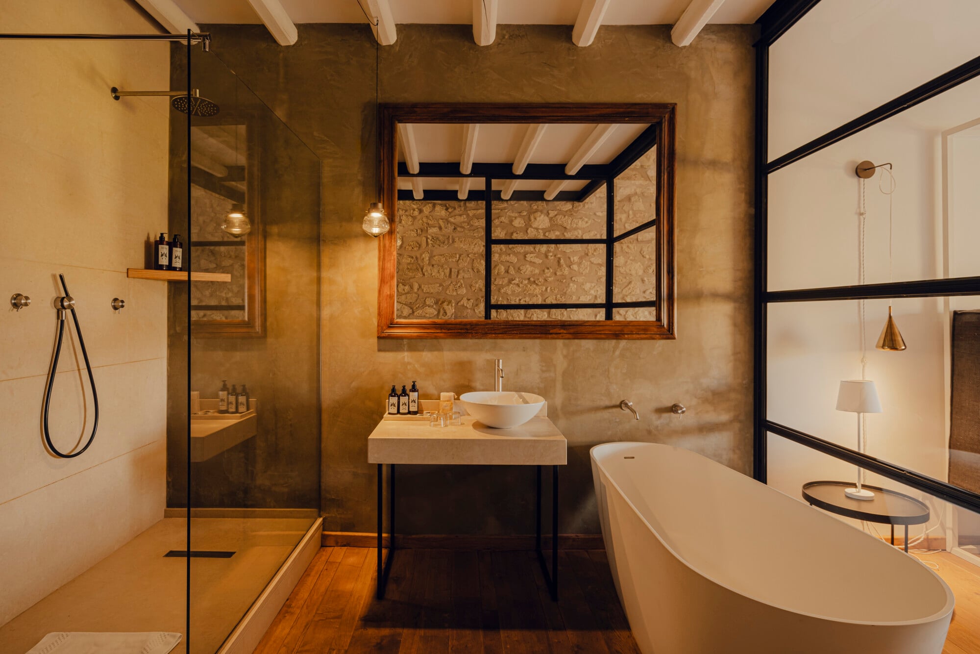 Susafa, Italy — Spa bathroom with travertine walls, floating stone vanity, freestanding soaking tub, walk-in shower, wooden beams, and framed mirror with black metal accents.