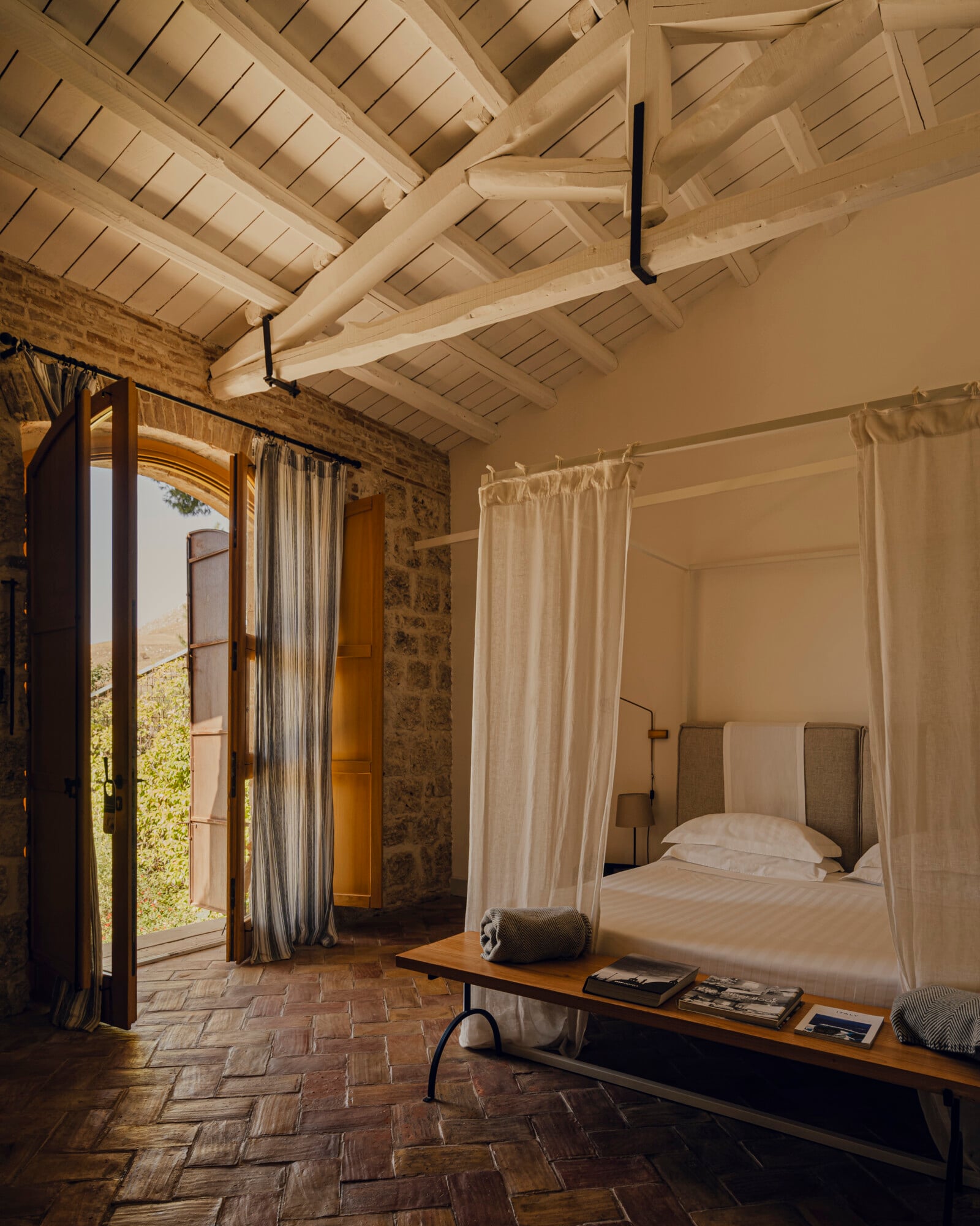 Susafa, Italy — Canopy bed in stone bedroom with exposed timber ceiling, terracotta floor, and open doors to landscape.