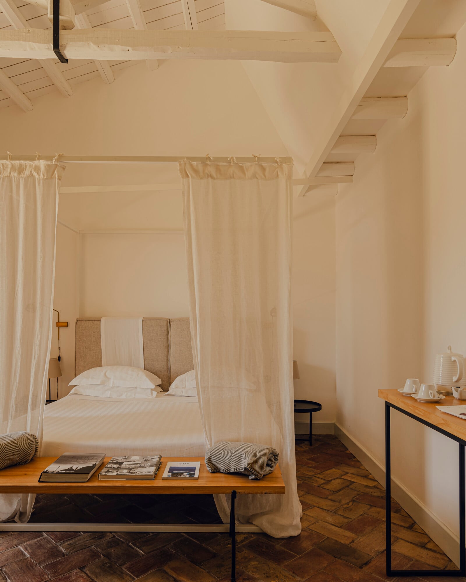 Susafa, Italy — Canopy bed with linen curtains in minimalist bedroom with exposed wooden ceiling beams and herringbone floor.