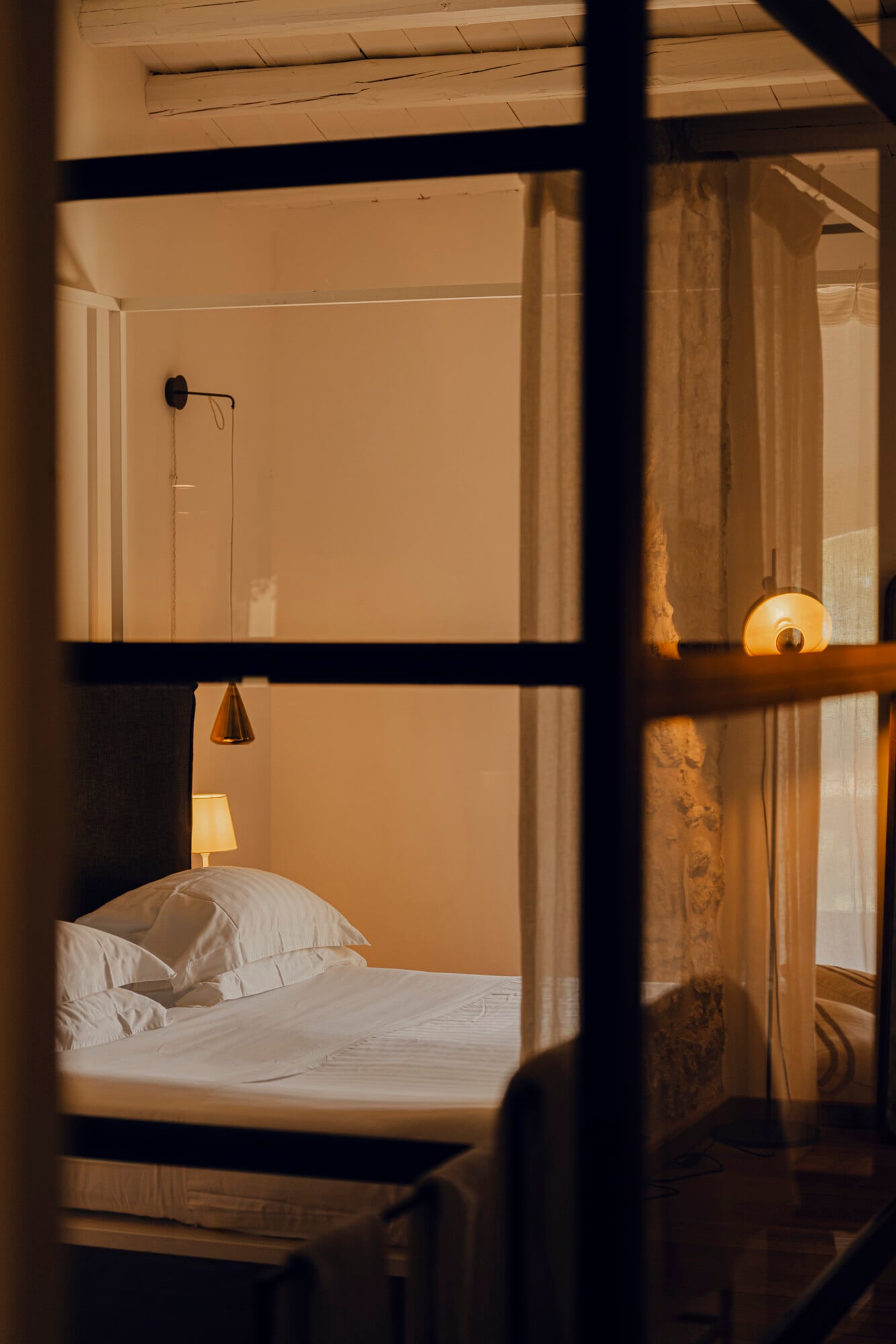 Susafa, Italy — Minimalist bedroom viewed through black-framed window, white bedding, warm ambient lighting, wooden architectural details, serene hospitality interior design.