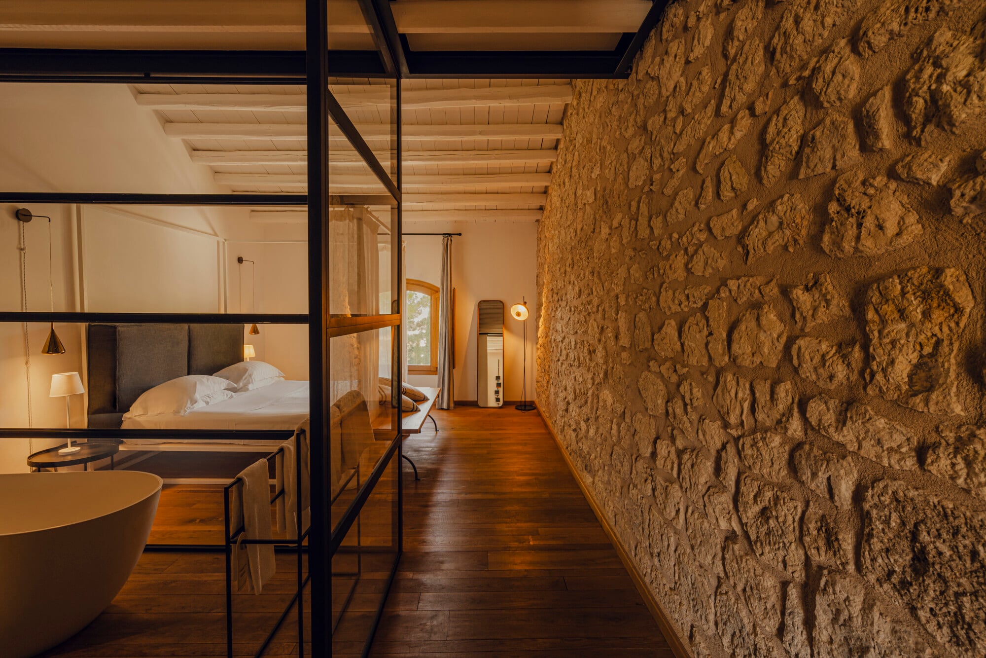 Susafa, Italy — Stone-walled bedroom with black-framed glass partition, wooden beams, warm ambient lighting, and minimalist furnishings overlooking countryside.