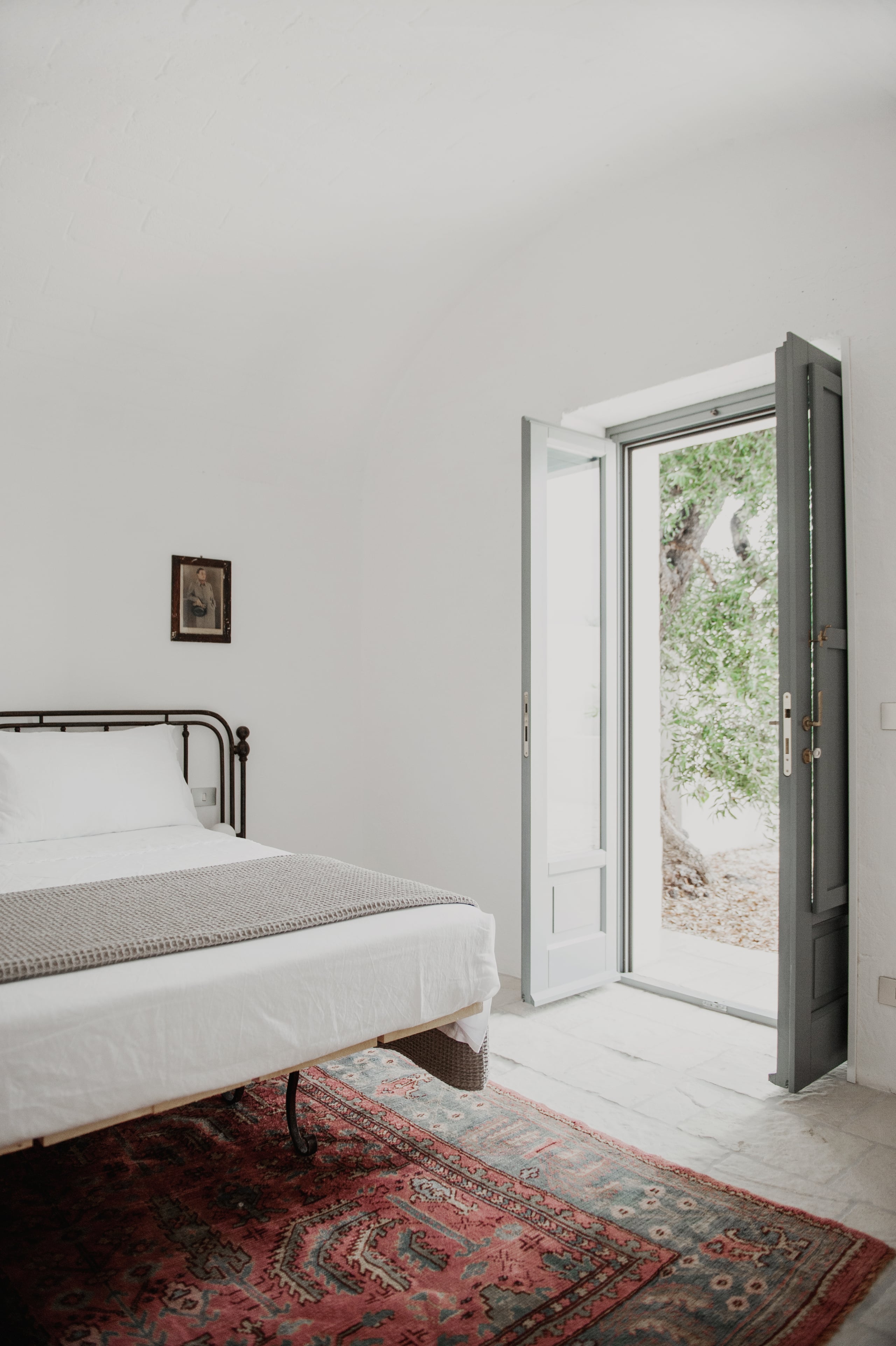 Masseria Moroseta, Europe — Minimalist bedroom with iron bed frame, vintage kilim rug, and open glass doors revealing stone courtyard with climbing ivy.