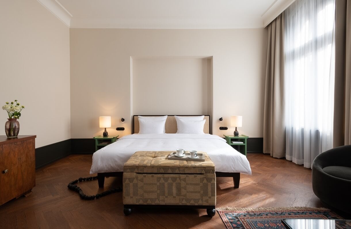 Château Royal, Europe — Minimalist luxury bedroom with white linens, woven headboard, green nightstands, warm wooden flooring, and floor-to-ceiling windows casting soft natural light.