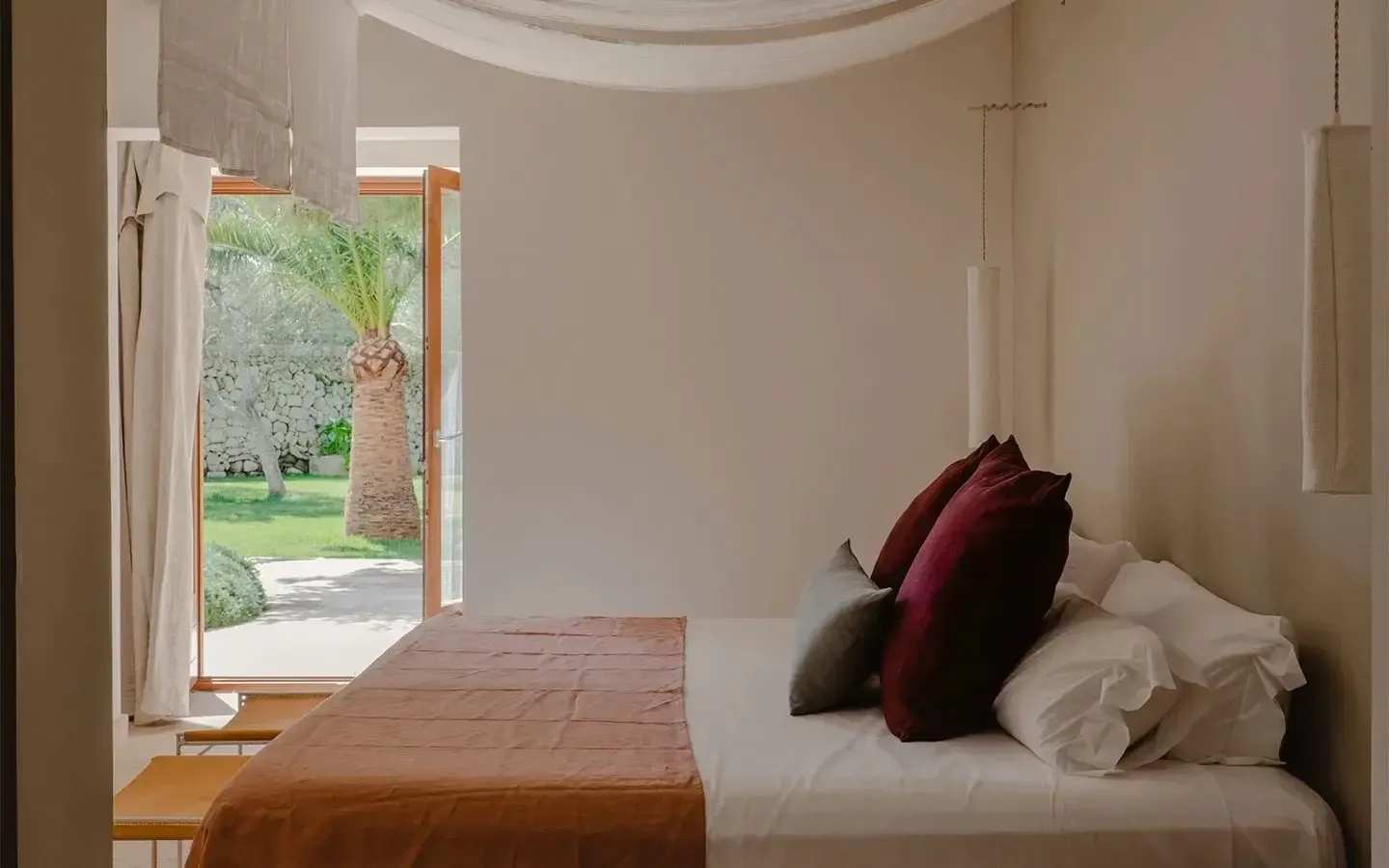 Amagatay, Balearic Islands — Minimalist bedroom with terracotta floor, cream walls, burgundy pillows, curved ceiling detail, and garden view through open doors.