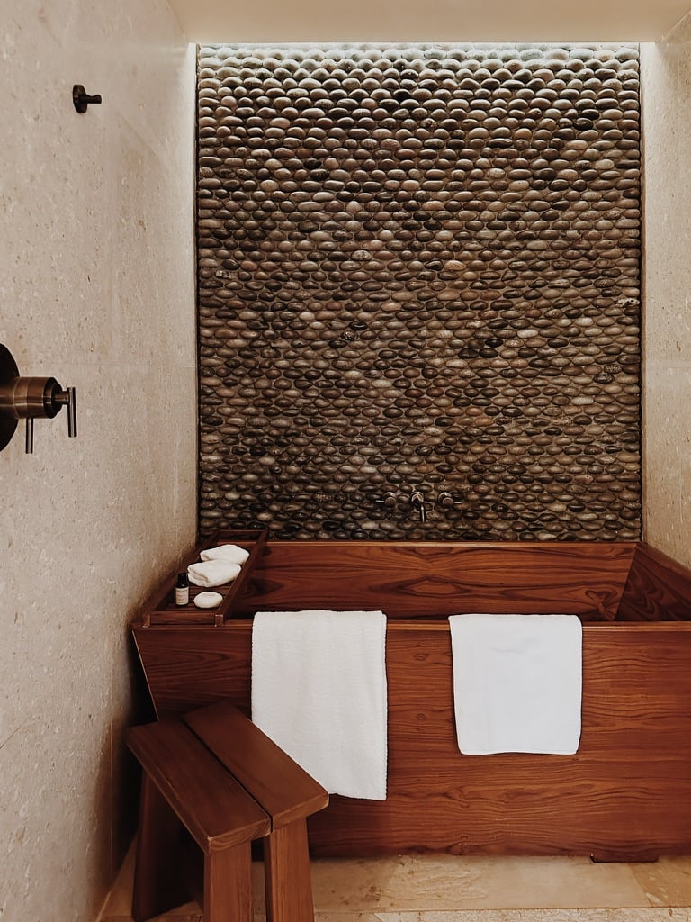 Nobu Los Cabos, Mexico — Wooden soaking tub with textured stone accent wall, white towels, minimalist spa bathroom with bronze fixtures.