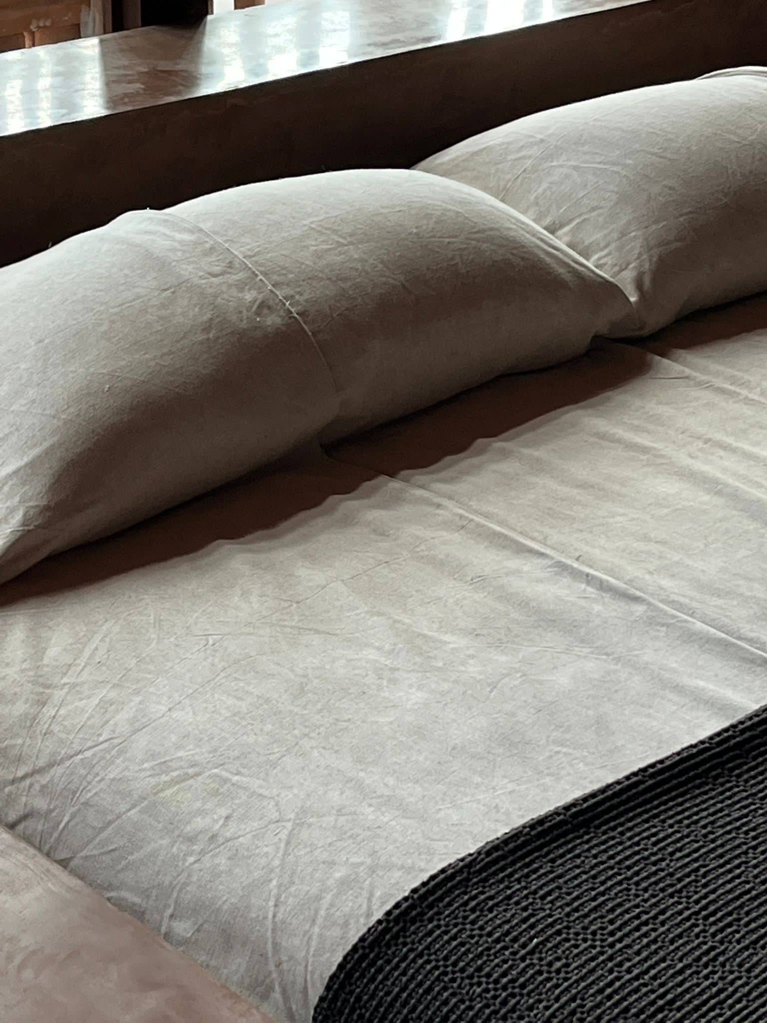 Further Hotel, Asia — Linen pillows and sheets on bed with dark wooden headboard, natural light casting shadows, minimalist luxury bedroom detail.