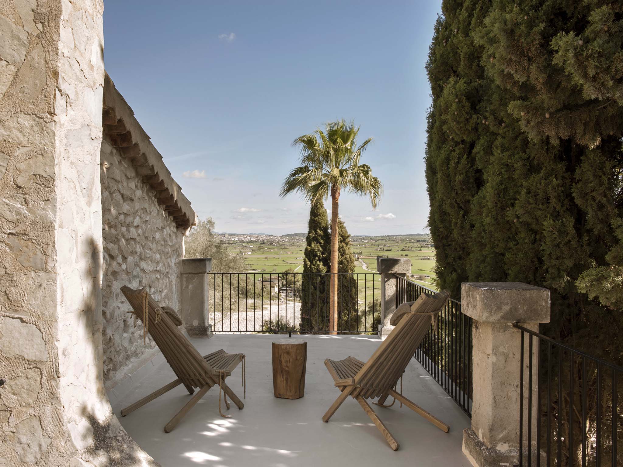 Finca Serena, Balearic Islands — Terraced stone villa overlooks green valley with palm tree, cypress hedges, wooden lounge chairs, and metal railing in Mediterranean light.