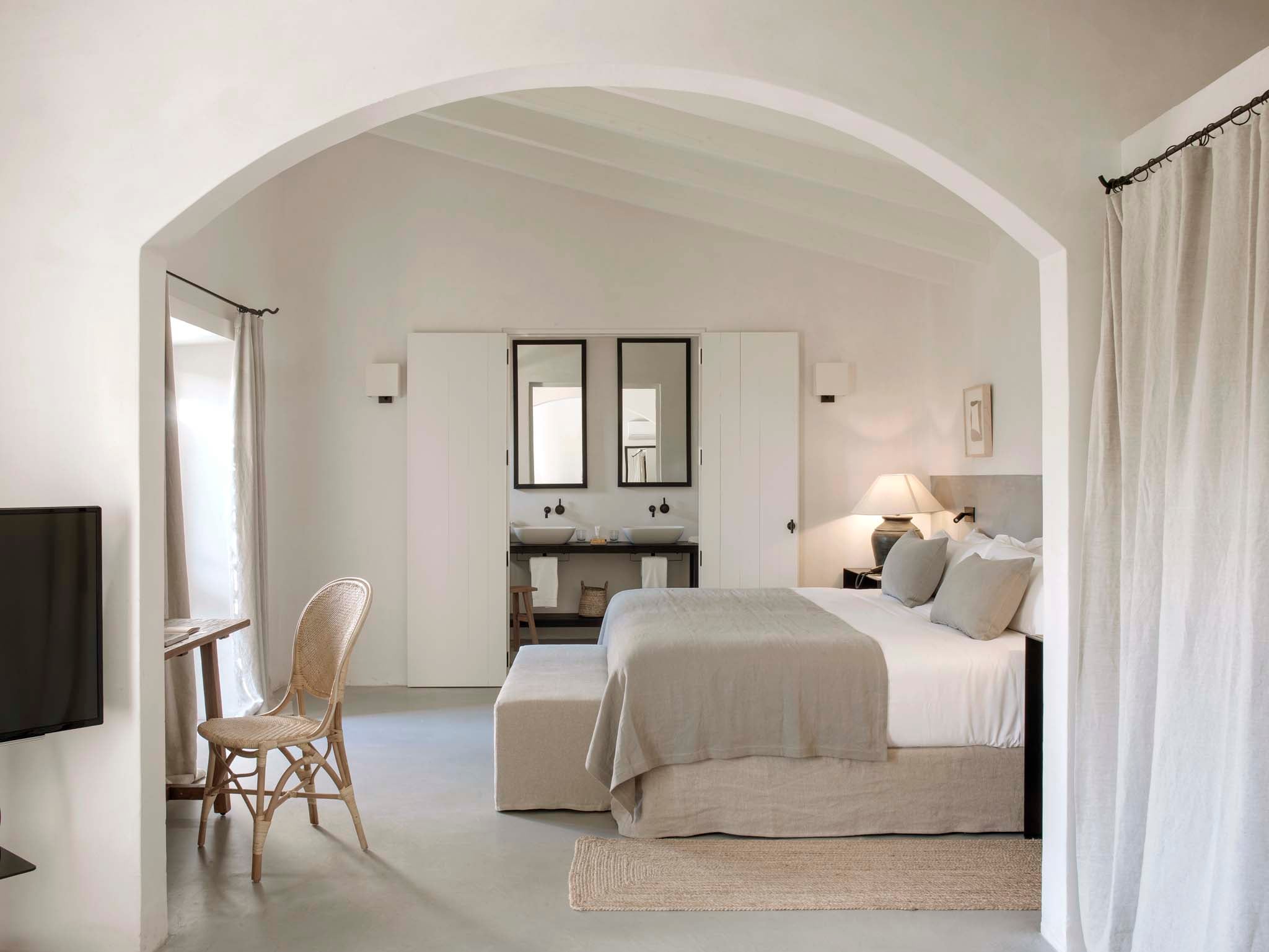 Finca Serena, Balearic Islands, Spain — Arched master bedroom suite with dual vanity, linen bedding, rattan chair, and ensuite bathroom.