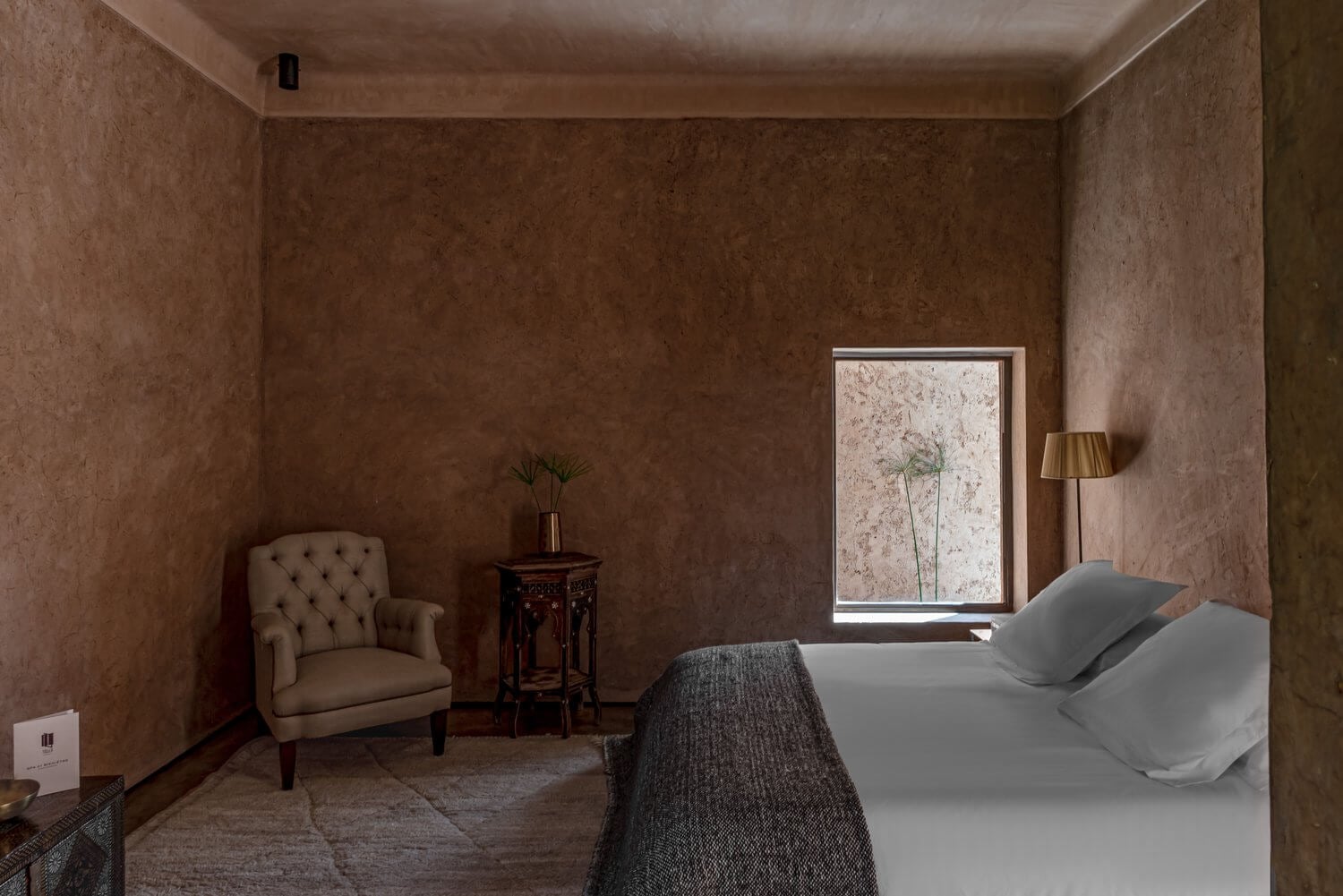 Villa D, Africa — Minimalist bedroom with terracotta textured walls, white bed, beige wingback chair, wooden side table, and large window with botanical artwork, warm ambient lighting.