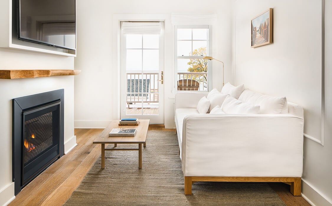 The Lake House, North America — Modern lakeside suite with fireplace, white upholstered bed, wooden furniture, and French doors opening to water views.