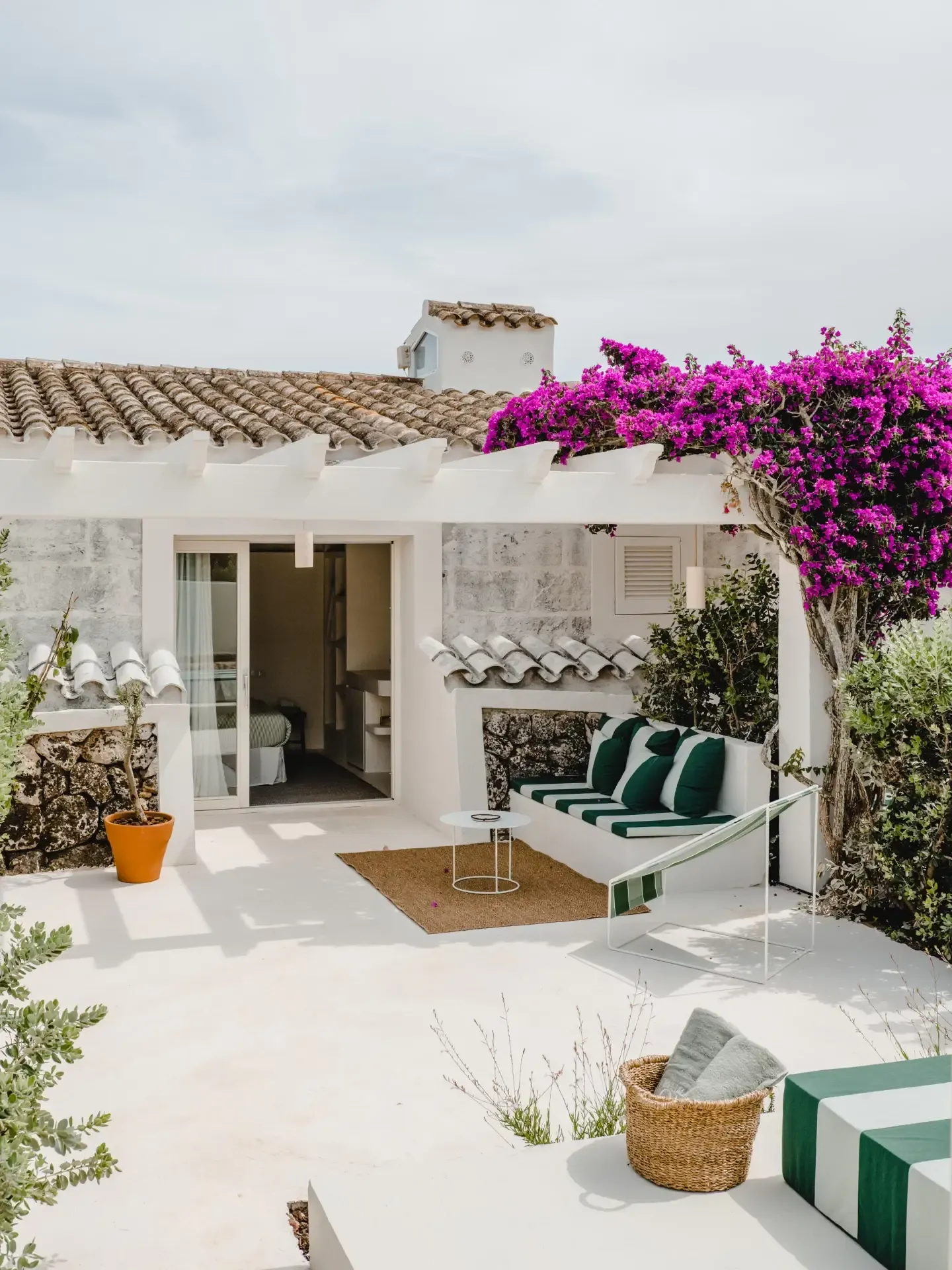 Morvedra Nou, Balearic Islands — Mediterranean courtyard with magenta bougainvillea, white-washed walls, terracotta roof tiles, and green-striped daybed seating.