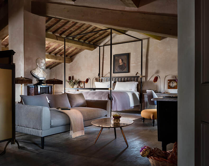 Castello di Reschio, Europe — Tuscan suite with canopy bed, mid-century furniture, exposed beam ceiling, stone floors, and classical portrait, blending medieval architecture with contemporary design.