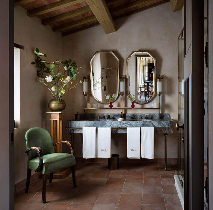 Castello di Reschio, Europe — Bathroom with dual marble vanity, octagonal mirrors, exposed timber ceiling, terracotta floors, green velvet chair, brass fixtures, and fresh flowers in gold vessel.
